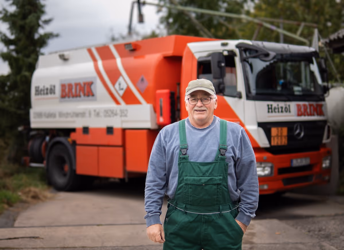 Älterer Mann mit grüner Arbeitslatzhose und Mütze steht vor einem orange-weißen Heizöl-Lieferwagen.