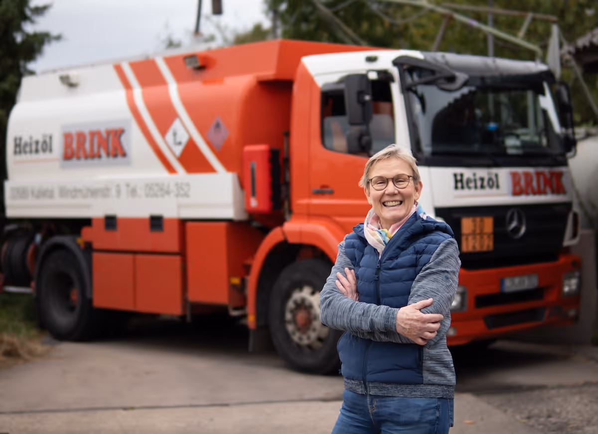Lächelnde ältere Frau mit Brille und blauer Weste steht vor einem orange-weißen Heizöl-Lieferwagen der Firma BRINK.