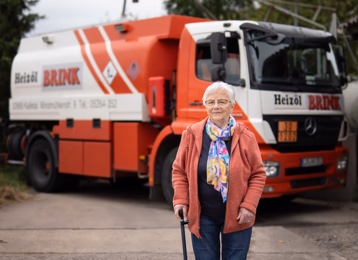 Ältere Frau mit buntem Schal und orangefarbener Jacke steht vor einem Heizöl-Lkw der Marke Brink.