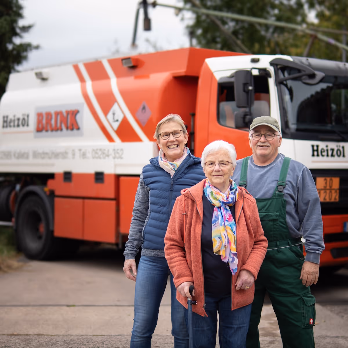 Drei ältere Personen lächelnd vor einem rot-weißen Heizöl-LKW mit der Aufschrift ‚BRINK‘.