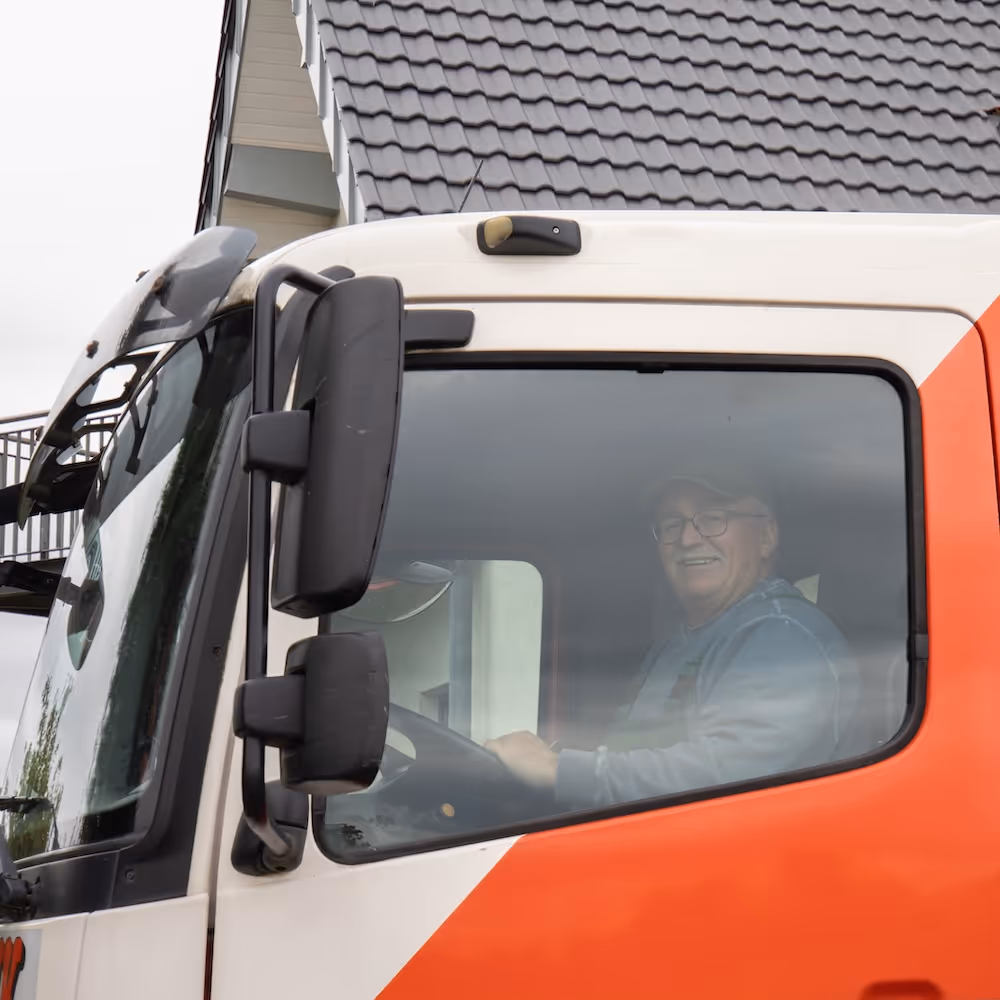 Älterer Mann mit Brille und Kappe sitzt lächelnd im Fahrersitz eines orange-weißen Lastwagens.