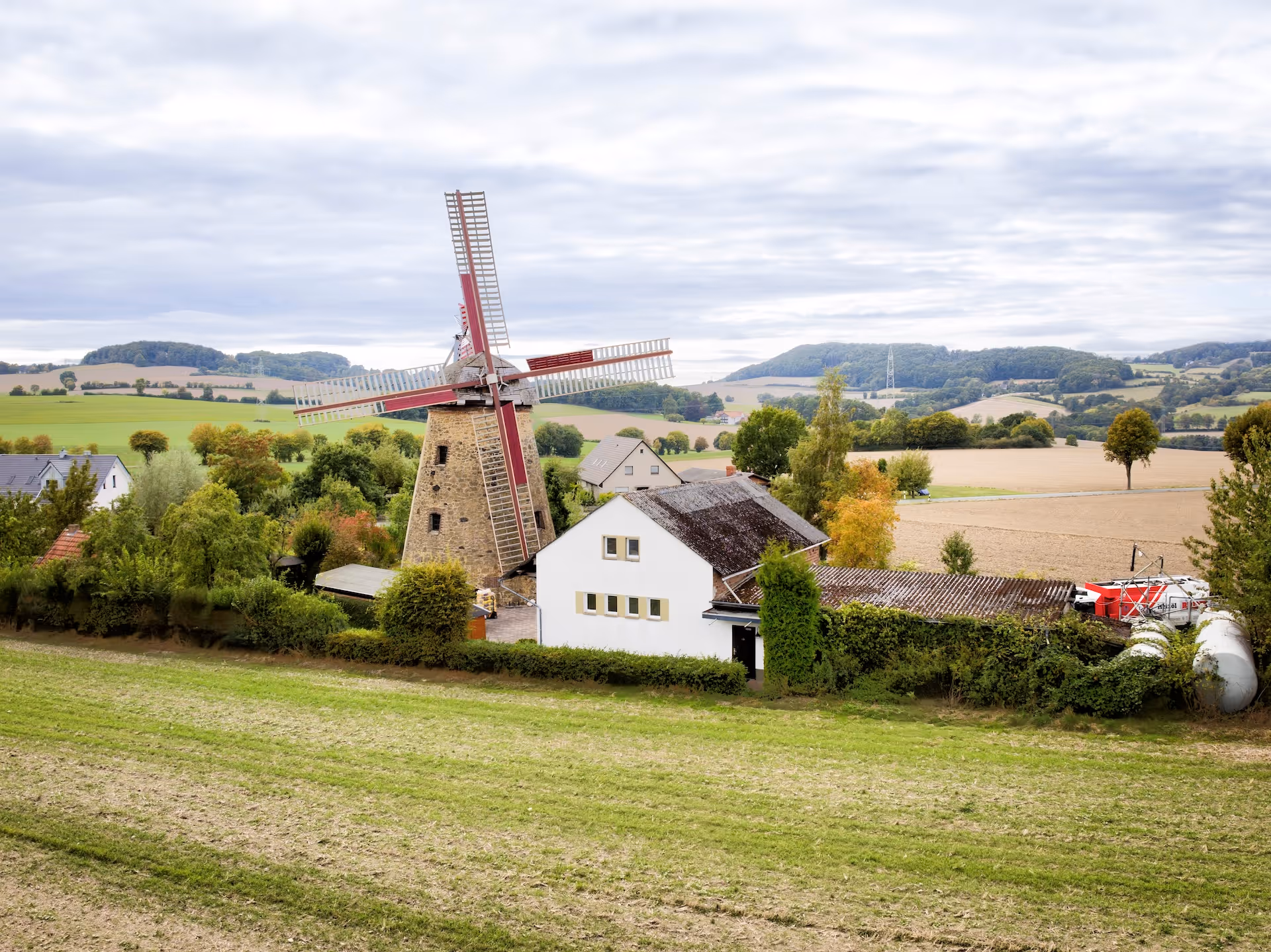 Eine historische Windmühle mit roten Flügeln steht neben einem weißen Haus in einer ländlichen Landschaft mit Feldern und bewaldeten Hügeln unter bewölktem Himmel.