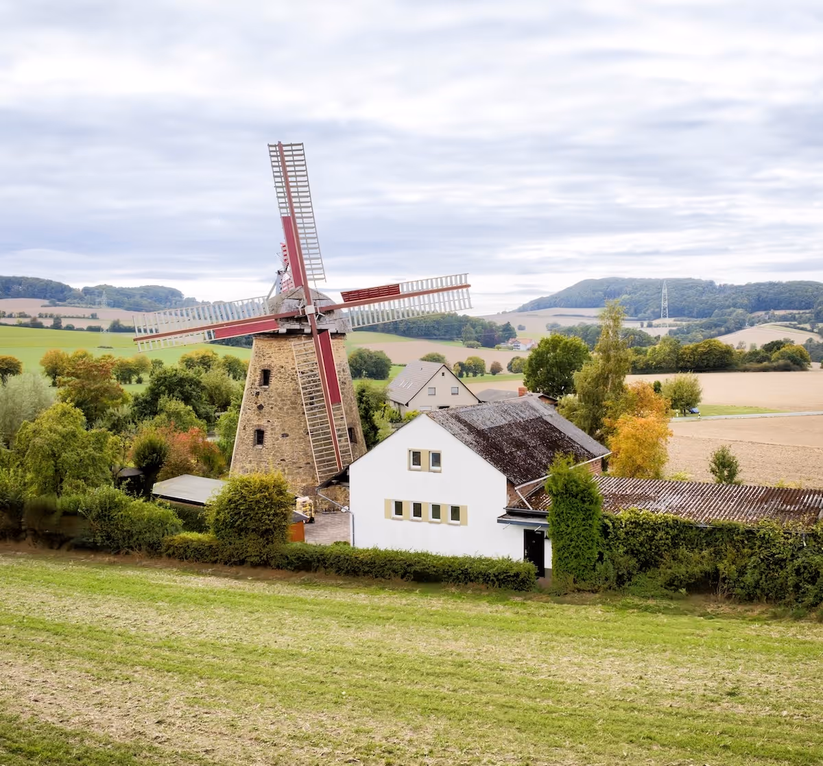 Steinwindmühle mit roten Flügeln neben einem weißen Haus auf grünem Feld mit Hügeln im Hintergrund.