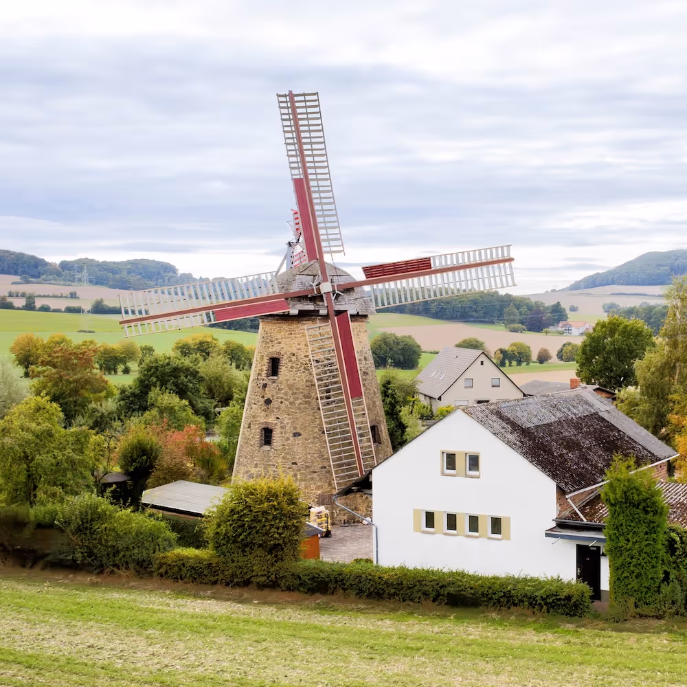 Steinmühle mit rot-weißen Flügeln neben einem weißen Haus in einer grünen ländlichen Landschaft mit Hügeln und bewölktem Himmel.