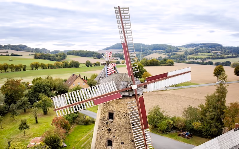 Luftaufnahme von zwei historischen Windmühlen vor Landschaft mit Feldern und Hügeln.