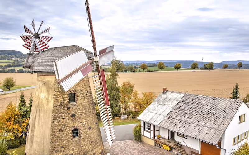 Eine Windmühle aus Stein neben einem Fachwerkhaus vor herbstlichen Feldern und bewölktem Himmel.